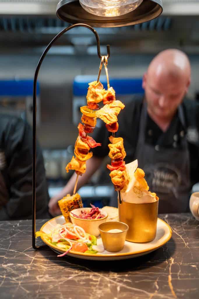 Cocktail presentation with skewered pineapple and fruit under heat lamp, served with golden drink on dark table.