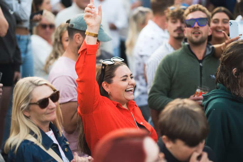 Crowd of spectators at an outdoor event, woman in red jacket with arm raised, smiling among other seated attendees.